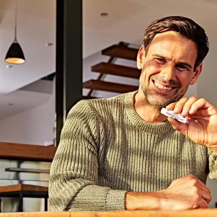 Homme souriant tenant un stick Forever Aloe Lips dans une ambiance chaleureuse. Hydratation des lèvres au quotidien.
